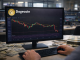 A trader’s hands operate a keyboard while monitoring a Dogecoin candlestick chart on a desktop monitor in a financial office setting.